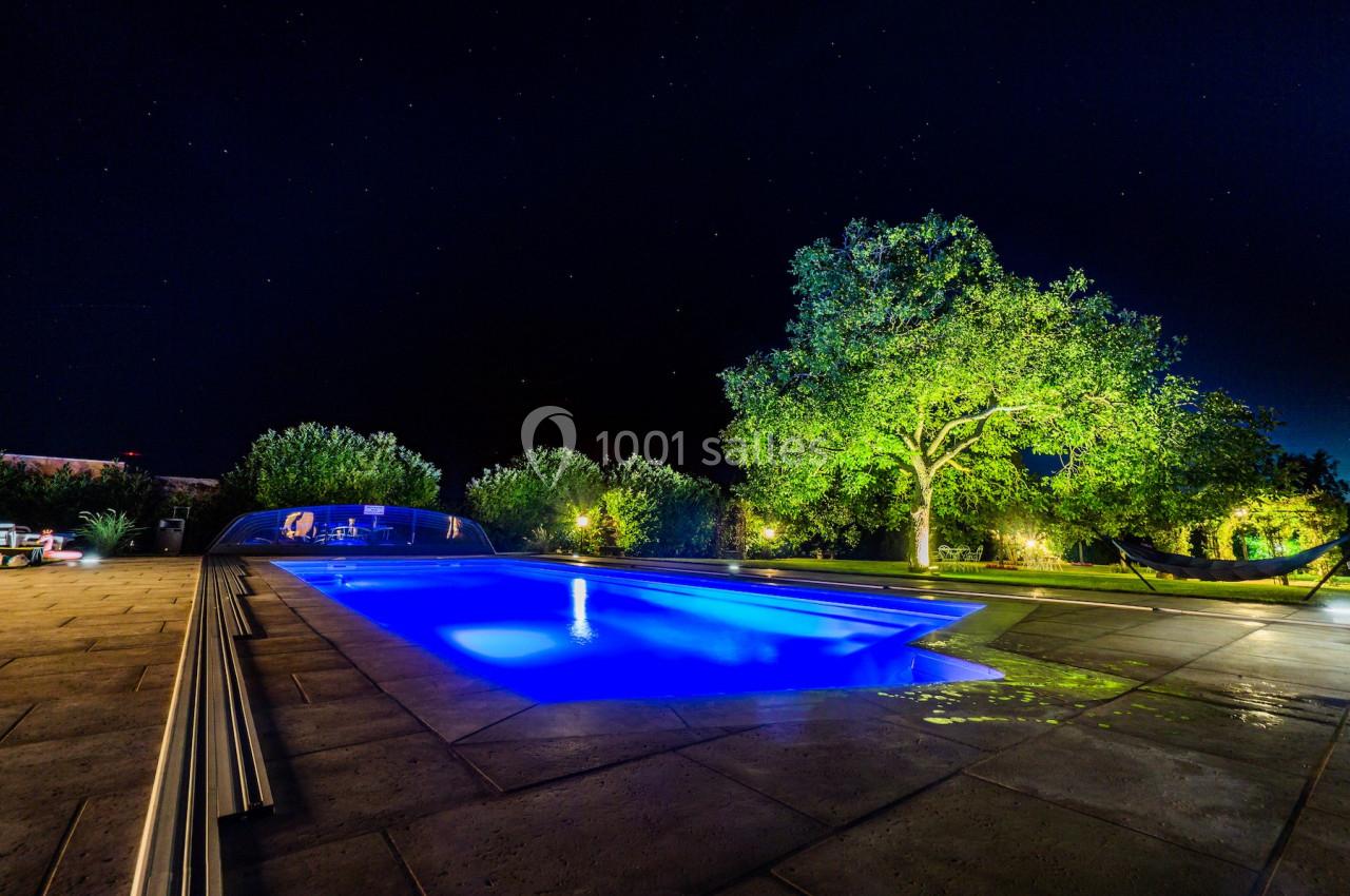 Piscine éclairée en bleu la nuit, entourée d'arbres et d'une terrasse en pierre avec un hamac à droite.