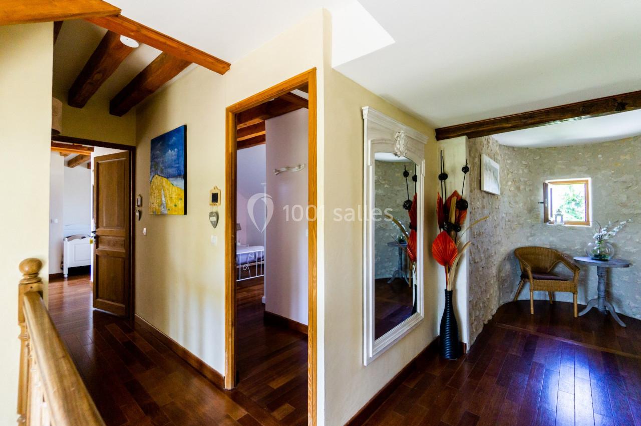 Vue d'un couloir intérieur avec parquet, poutres apparentes, miroir mural et décoration florale près d'une fenêtre.