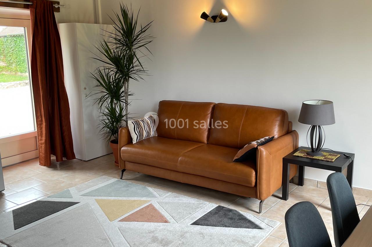 Salon lumineux avec canapé en cuir marron, tapis géométrique, table en bois, plantes et poutres apparentes au plafond.