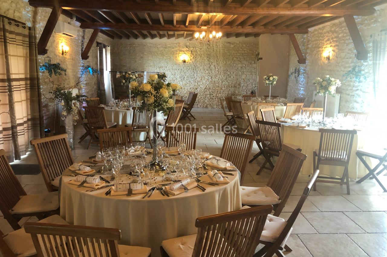 Salle de réception avec tables rondes dressées, chaises en bois et décoration florale dans un cadre en pierre apparente.