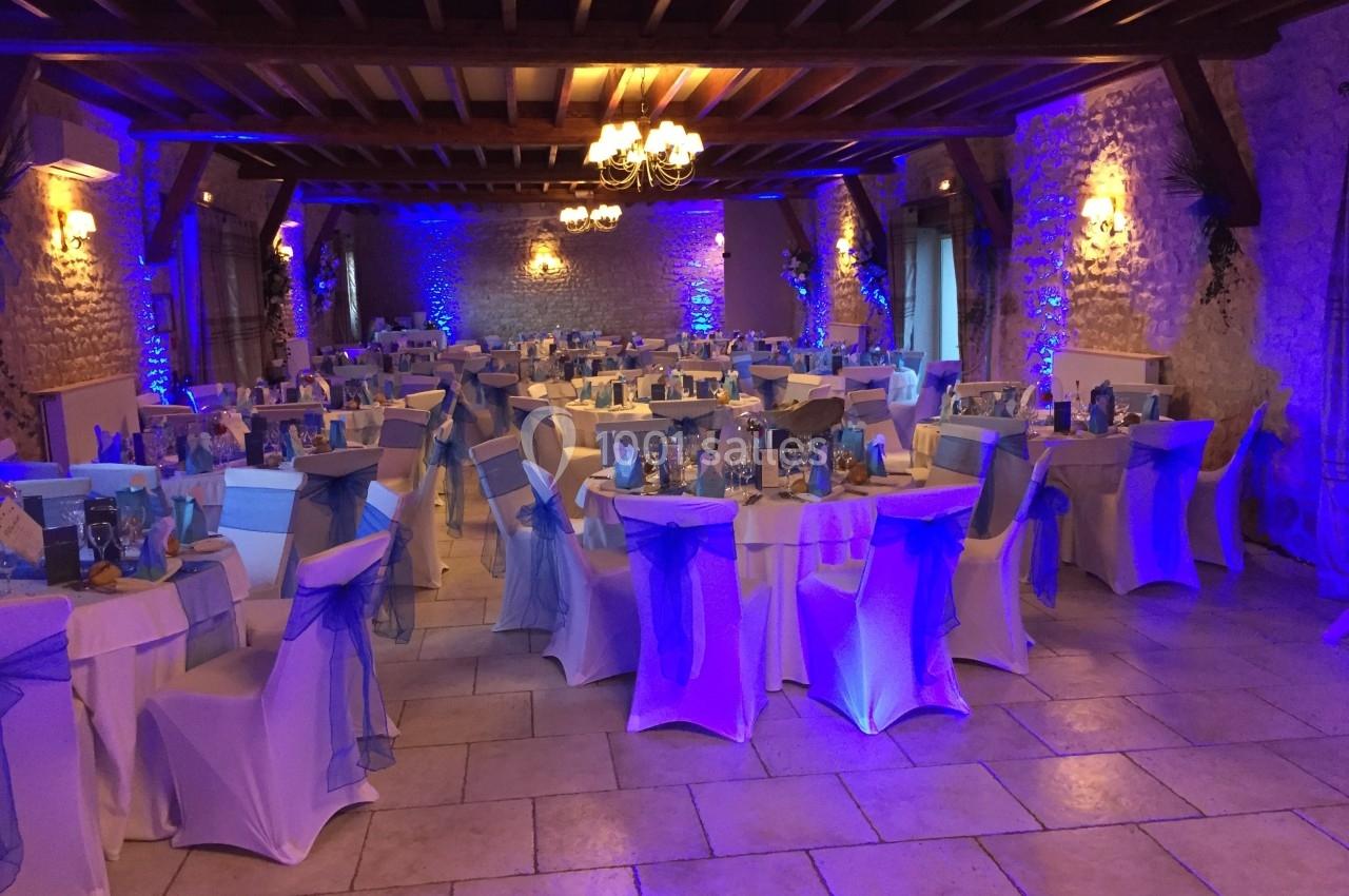 Salle de réception décorée pour un événement, avec des tables rondes, nappes blanches et éclairage bleu.