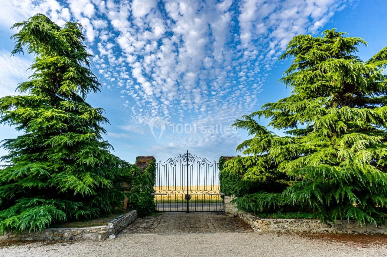 Portail en fer forgé entouré de grands arbres verts, sous un ciel bleu parsemé de nuages.