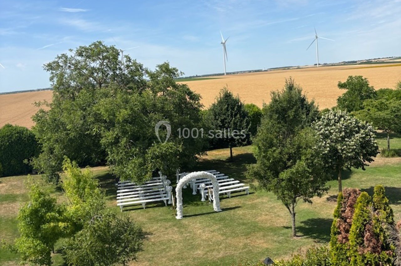 Vue d'un jardin avec des bancs blancs disposés devant une arche décorée, entouré d'arbres et de champs avec des éoliennes au…