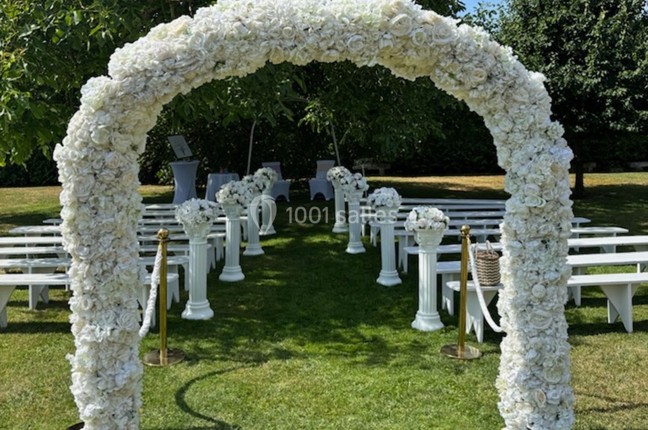 Arche de fleurs blanches menant à une allée bordée de bancs et de colonnes fleuries dans un jardin verdoyant.