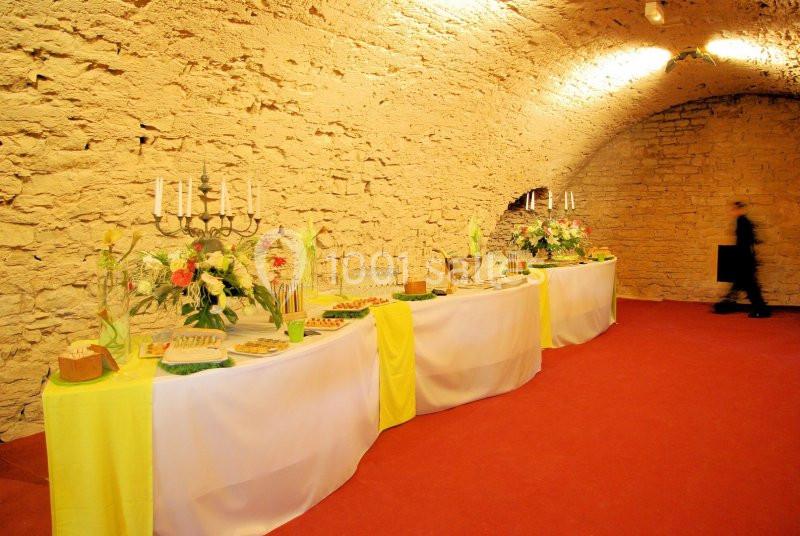 Table de buffet décorée avec nappes blanches et jaunes, fleurs et chandeliers, dans une salle voûtée en pierre.