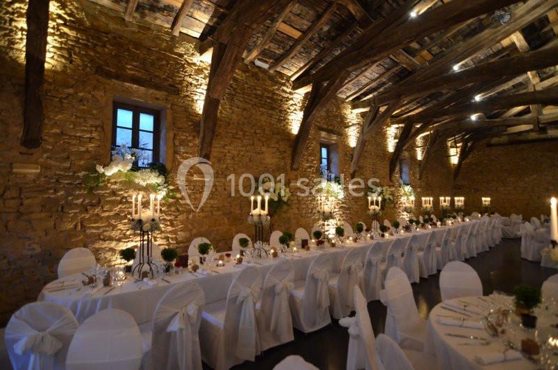 Salle de réception en pierre avec poutres apparentes, tables décorées et chaises blanches alignées pour un événement.