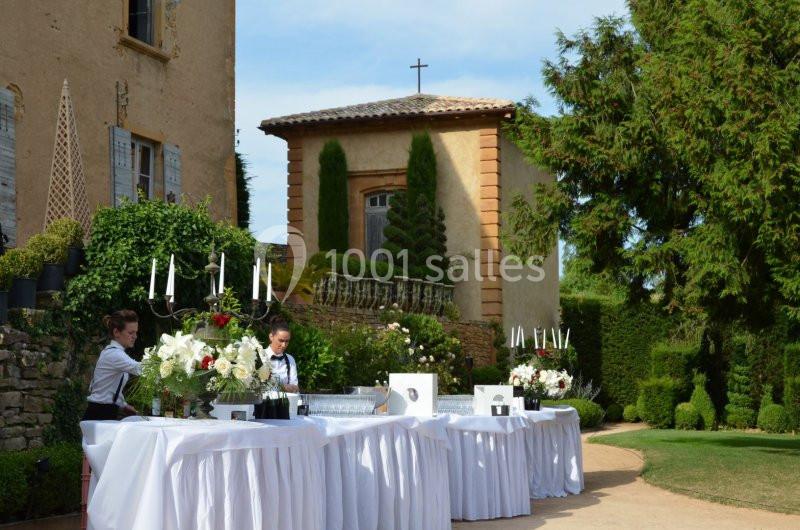 Tables dressées avec nappes blanches, décor floral et bougies, dans un jardin près d'une chapelle en pierre.