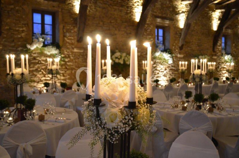 Salle de réception en pierre éclairée, décorée de chandeliers, fleurs blanches et tables dressées pour un événement.