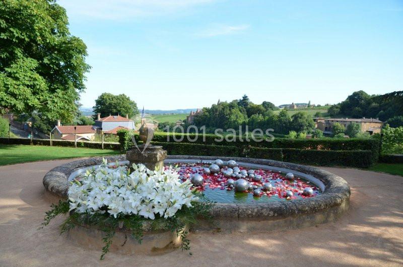 Bassin circulaire décoré de fleurs blanches et de boules argentées, entouré de verdure et offrant une vue sur la campagne.