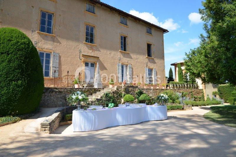 Table dressée en extérieur devant une grande bâtisse en pierre, entourée de végétation et d'un jardin aménagé.