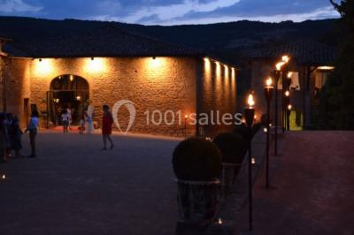 Cour éclairée au crépuscule avec des bâtiments en pierre, des torches allumées et des personnes en arrière-plan.