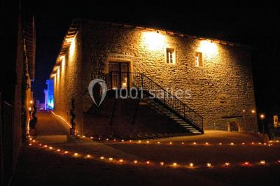 Cour éclairée au crépuscule avec des bâtiments en pierre, des torches allumées et des personnes en arrière-plan.