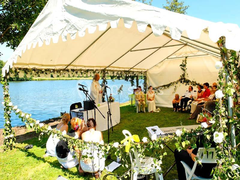 Cérémonie en plein air sous une tente décorée de feuillage, près d'un lac, avec des invités assis.