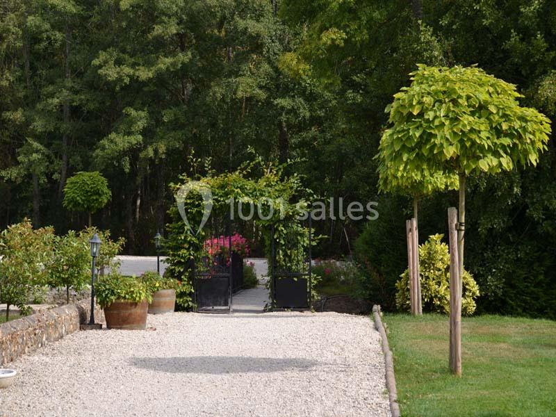 Allée gravillonnée bordée de verdure menant à un portail noir entouré de plantes et d'arbustes.