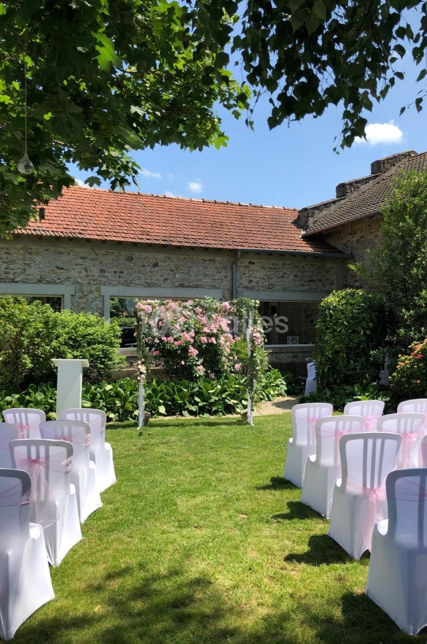 Chaises blanches décorées de rubans roses alignées devant une arche fleurie dans un jardin ensoleillé.