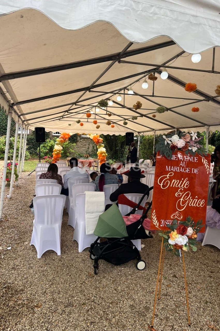 Cérémonie de mariage en extérieur sous une tente, avec des chaises blanches et une arche décorée de fleurs orange.