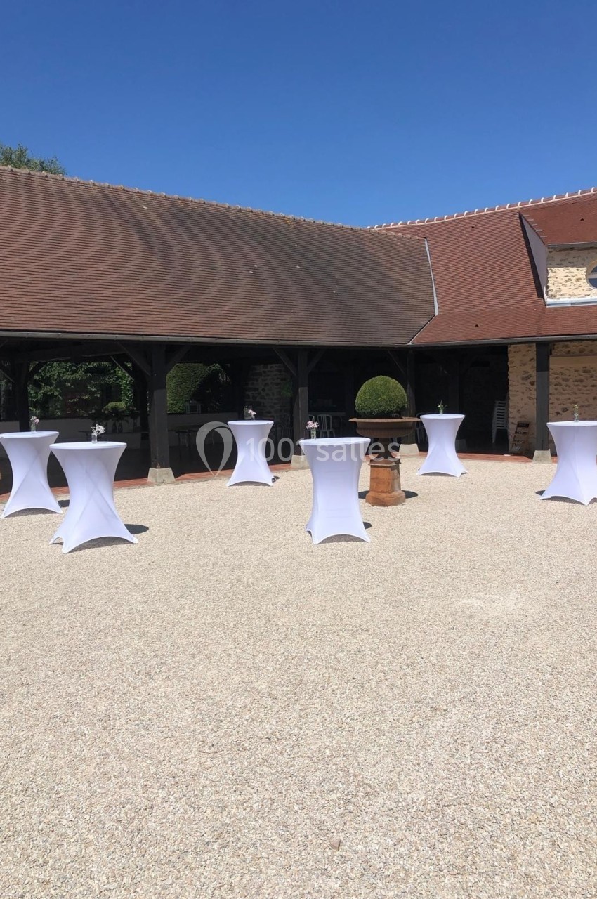 Tables hautes recouvertes de nappes blanches disposées en extérieur sur une cour pavée devant des bâtiments en pierre.