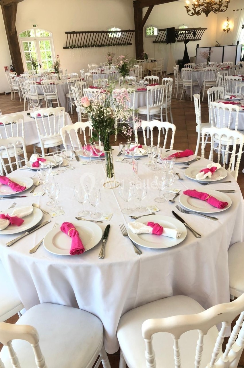 Salle de réception décorée avec des tables rondes, nappes blanches, serviettes roses et centres de table fleuris.