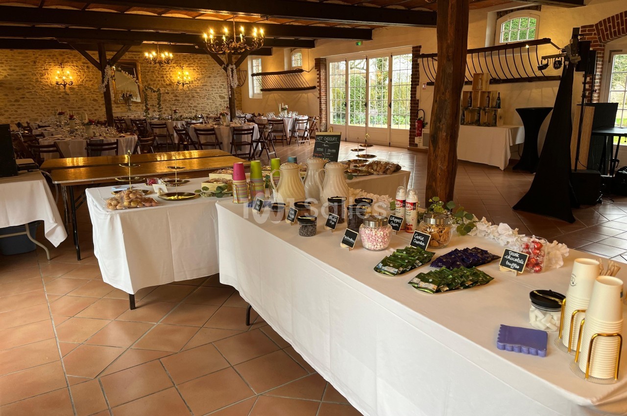 Tables dressées avec des buffets de nourriture et boissons dans une salle de réception lumineuse au style rustique.