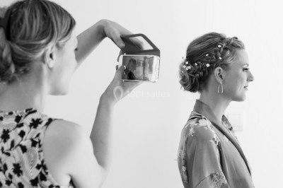 Femme en robe en dentelle blanche, coiffée d'une tresse et d'une fleur, dans un intérieur lumineux.