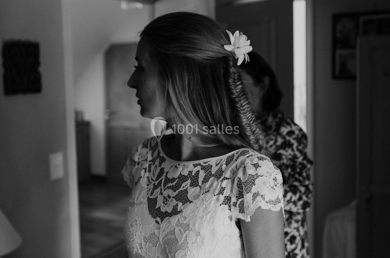 Femme en robe en dentelle blanche, coiffée d'une tresse et d'une fleur, dans un intérieur lumineux.