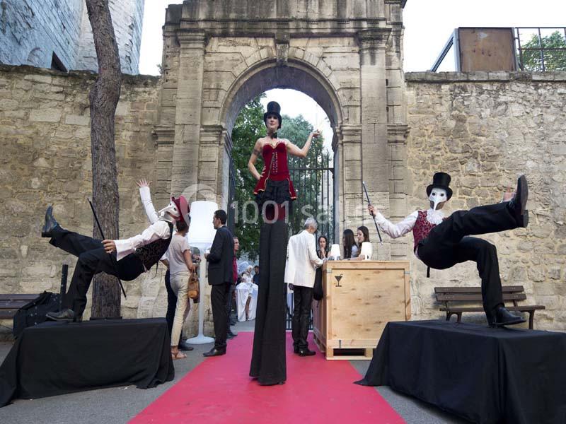 Deux artistes en costume, l'un sur échasses et l'autre masqué, posent sur un tapis rouge devant une arche en pierre.
