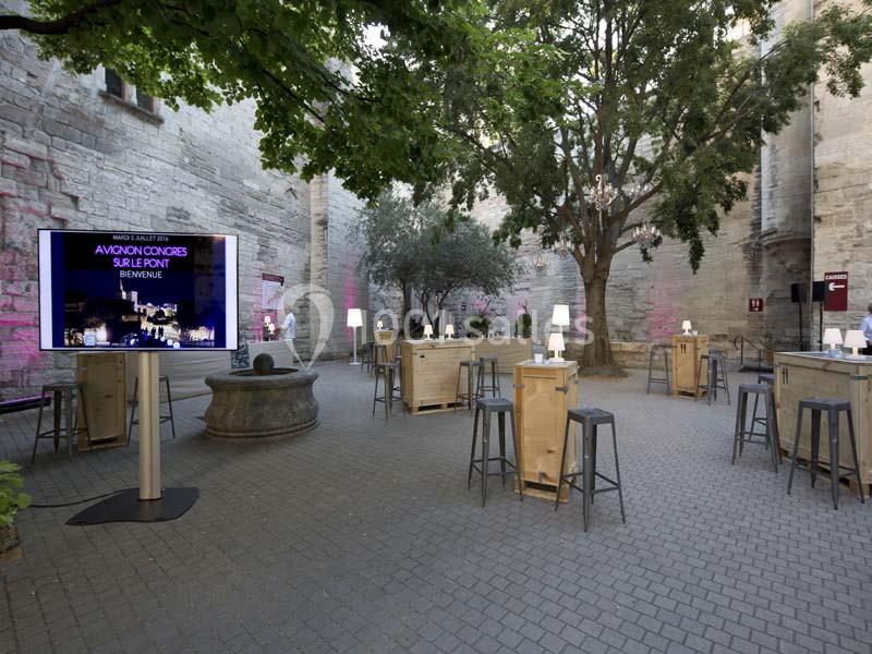 Cour intérieure pavée aménagée avec des tables hautes, un écran affichant ’Avignon Congrès’ et des arbres éclairés.
