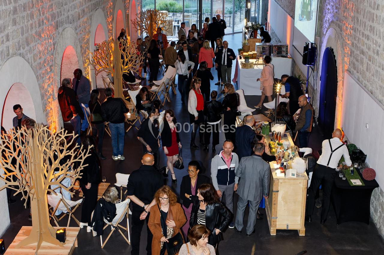 Salle animée avec des invités discutant autour de stands, dans un espace lumineux aux murs en pierre et grandes fenêtres.