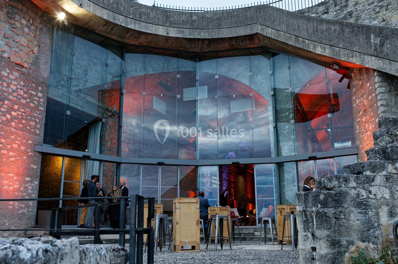 Façade vitrée d'un bâtiment moderne avec éclairages rouges, intégrée à des murs en pierre ancienne.