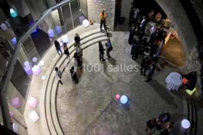 Façade vitrée d'un bâtiment moderne avec éclairages rouges, intégrée à des murs en pierre ancienne.