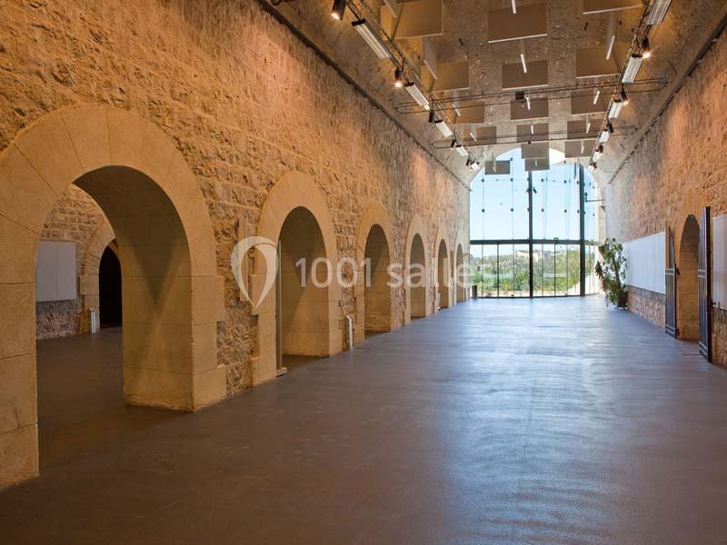 Intérieur d'une grande salle en pierre avec des arches, éclairée par des spots et une baie vitrée donnant sur l'extérieur.