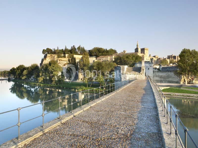 Vue d'un pont en pierre menant à une ville fortifiée avec des arbres et un fleuve reflétant le paysage.