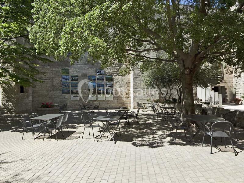 Terrasse ombragée avec des tables et des chaises en métal, entourée de murs en pierre et d'arbres.