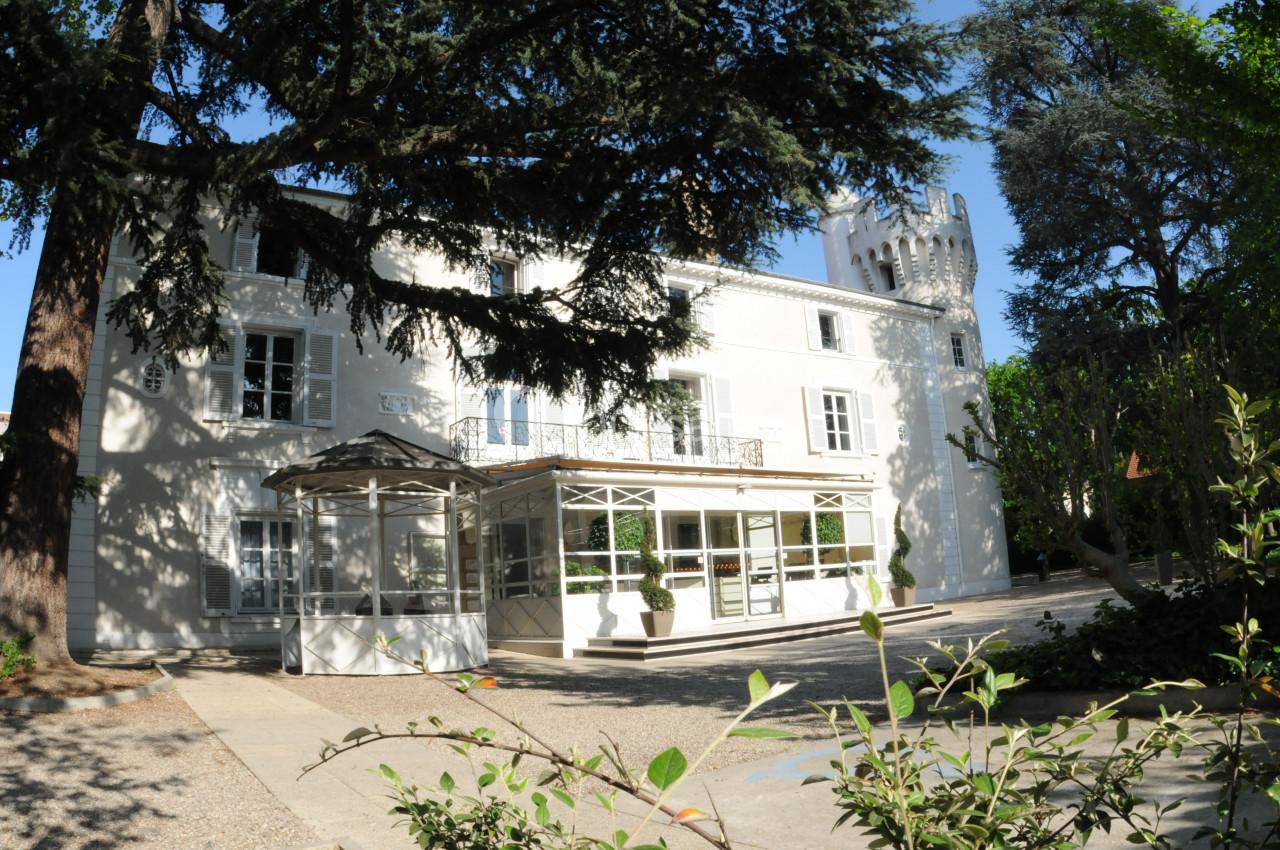 Façade d'un bâtiment blanc entouré d'arbres, avec une verrière et une cour en gravier au premier plan.