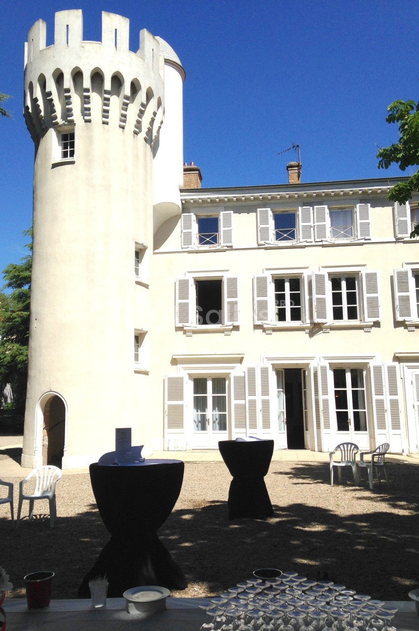 Tour blanche et bâtiment avec volets, tables hautes et chaises disposées dans une cour ensoleillée.