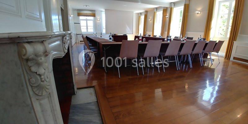 Salle de réunion lumineuse avec une grande table rectangulaire entourée de chaises, parquet en bois et cheminée en marbre.