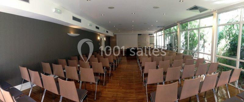 Salle de conférence lumineuse avec des rangées de chaises marron et une table au fond, entourée de grandes fenêtres.