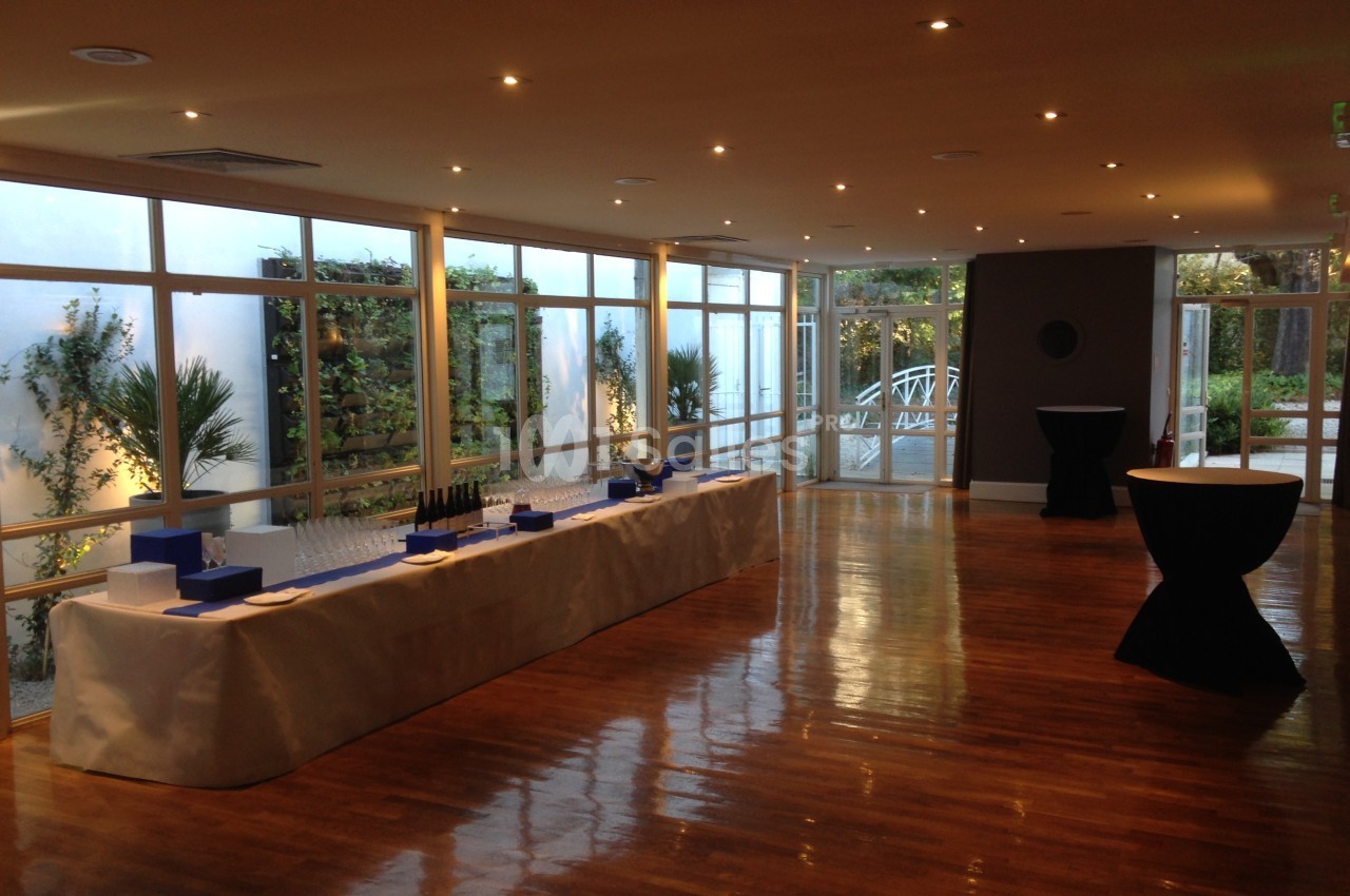 Salle lumineuse avec parquet, grandes baies vitrées, table dressée pour un événement et vue sur un jardin extérieur.