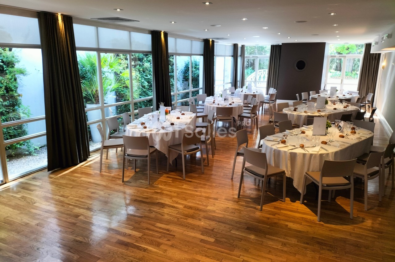 Salle lumineuse avec parquet, tables rondes dressées pour un repas, grandes baies vitrées donnant sur un jardin.