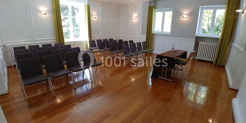 Salle lumineuse avec parquet, rangées de chaises noires, table en bois et grandes fenêtres avec rideaux verts.
