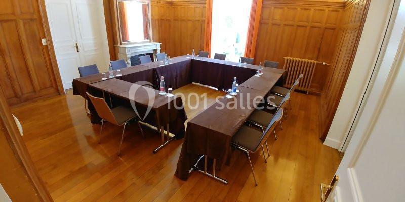 Salle de réunion avec tables disposées en U, chaises, bouteilles d'eau et lumière naturelle entrant par les fenêtres.