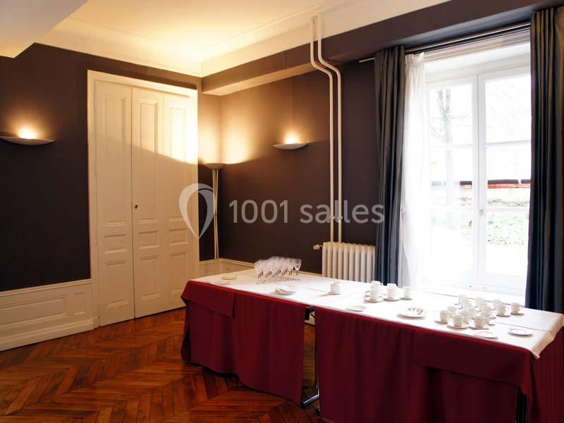 Salle lumineuse avec parquet en bois, table couverte de nappes rouges, et vaisselle disposée près d'une fenêtre.
