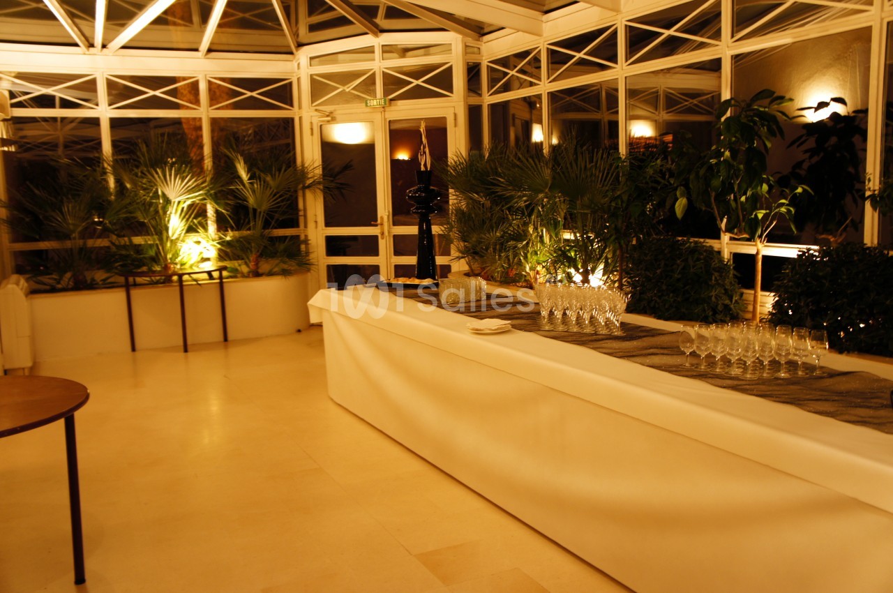 Salle lumineuse avec une grande table couverte d'une nappe blanche, verres alignés et plantes en arrière-plan.