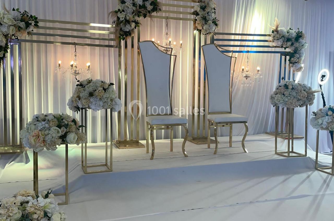 Deux chaises élégantes sur une scène décorée de fleurs blanches et dorées, avec un fond en tissu plissé.