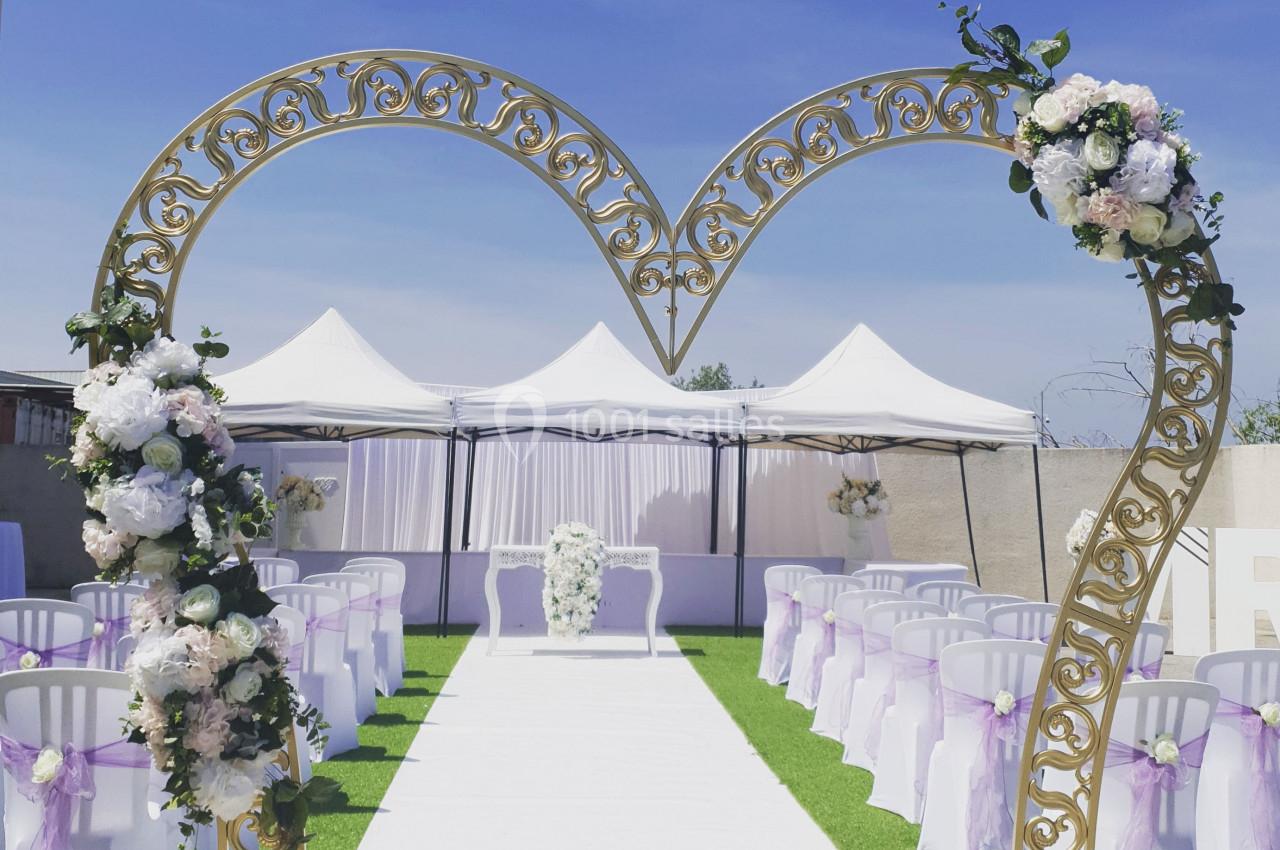 Arche dorée en forme de cœur décorée de fleurs blanches, encadrant une allée blanche menant à un autel extérieur.