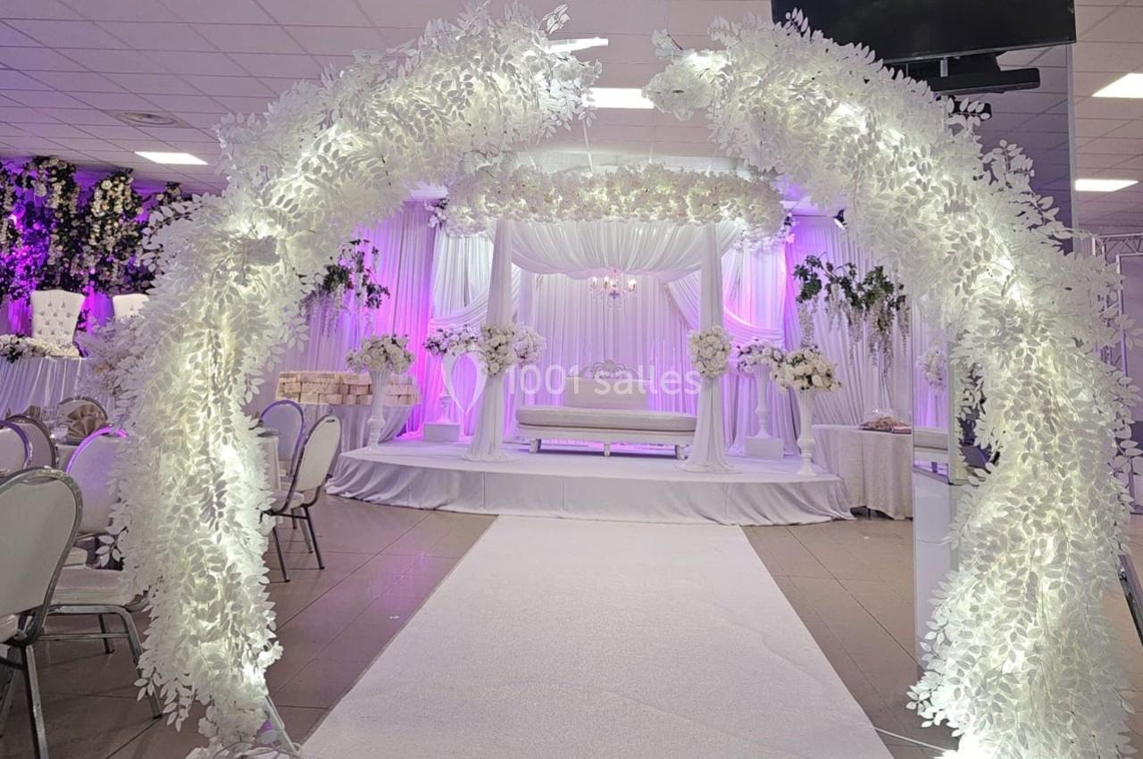 Arche florale blanche illuminée menant à une scène décorée de fleurs et de rideaux blancs dans une salle de réception.