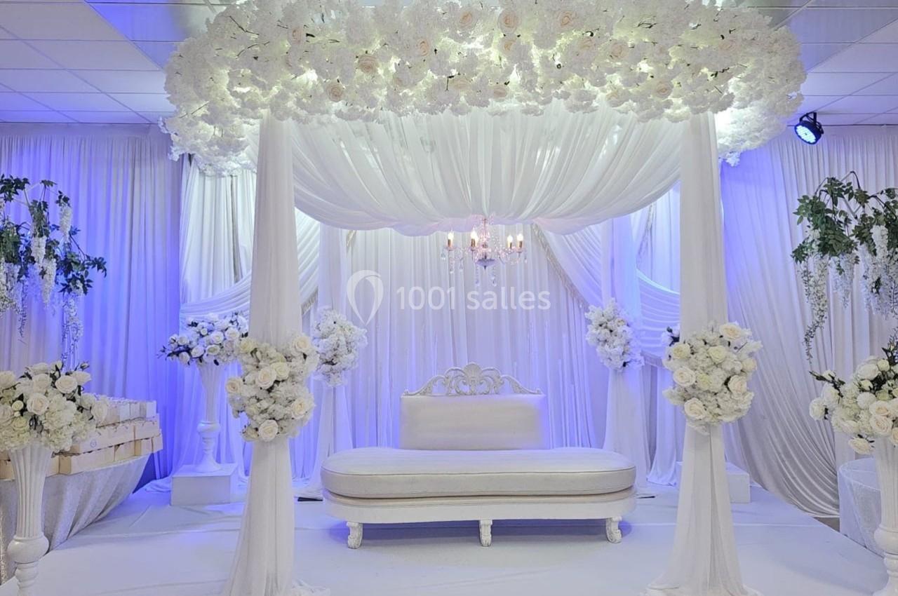 Décor de mariage élégant avec un canapé blanc sous une arche drapée de tissu blanc et ornée de fleurs.