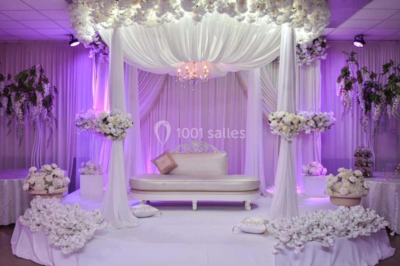 Décor de mariage avec canapé blanc sous une arche drapée de tissu blanc, entourée de fleurs et éclairée en violet.