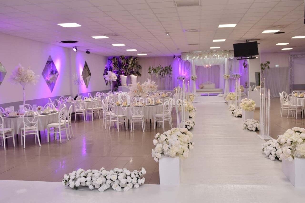 Salle de réception décorée en blanc et violet avec tables dressées, allée centrale et fleurs blanches.
