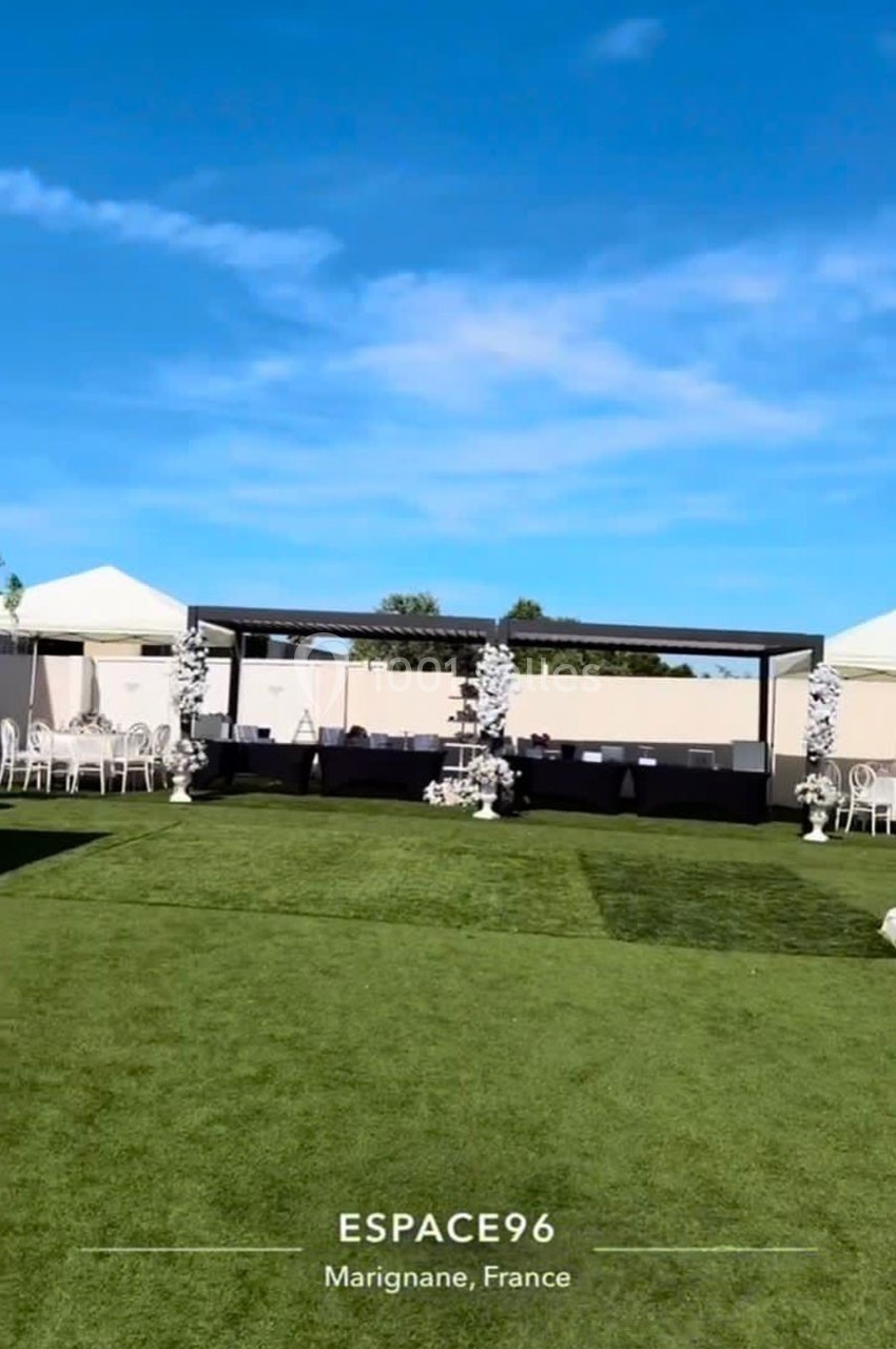 Vue d'un espace extérieur aménagé avec pelouse, tentes blanches, chaises et décoration florale sous un ciel bleu.